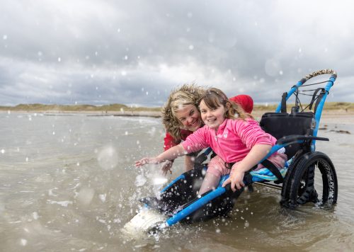 A mother and her daughter in the sea at the beach, they are enjoying the water together and splashing around. They are both looking at the camera and smiling. The daughter is a wheelchair user.