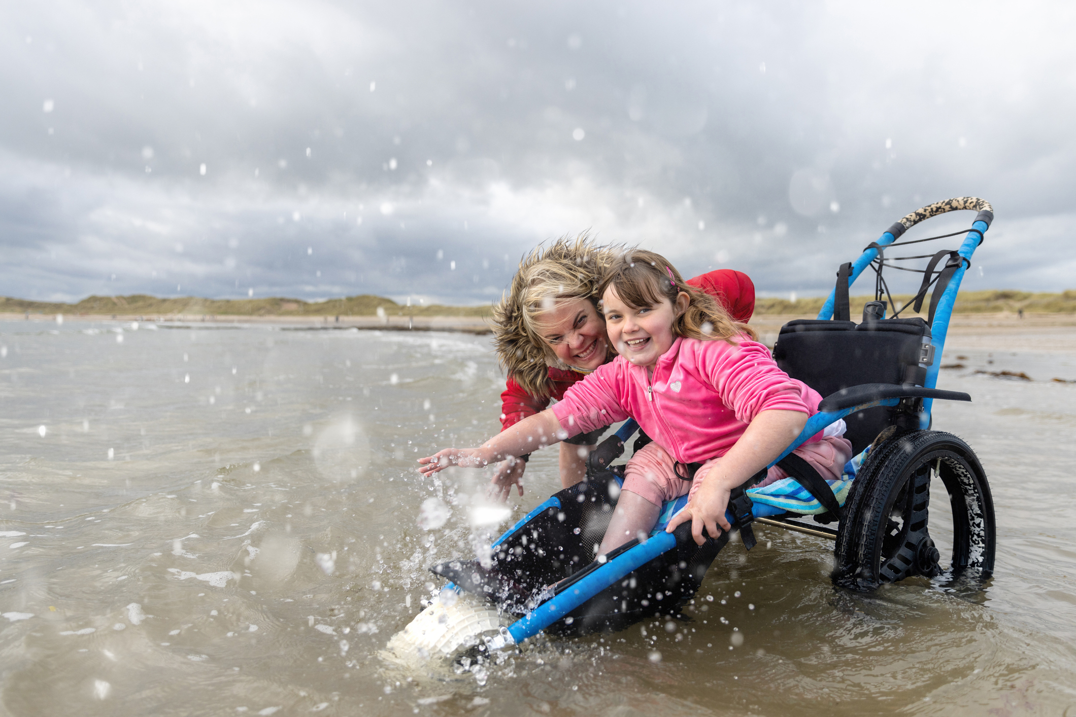 A mother and her daughter in the sea at the beach, they are enjoying the water together and splashing around. They are both looking at the camera and smiling. The daughter is a wheelchair user.