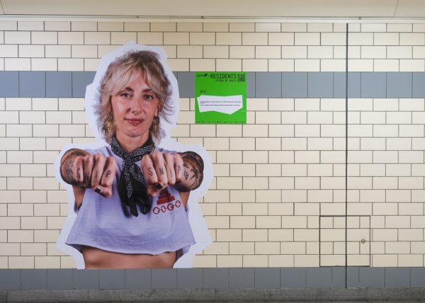 A photo of a person wearing a white t-shirt with attoos on their fingers and arms. The photo is on a tiled wall in a train station.