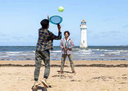 Two people playing tennis ona beach - there is a lighthouse in the background.