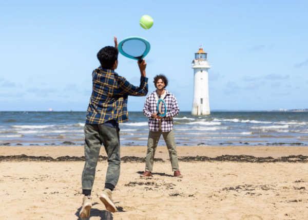 Two people playing tennis ona beach - there is a lighthouse in the background.