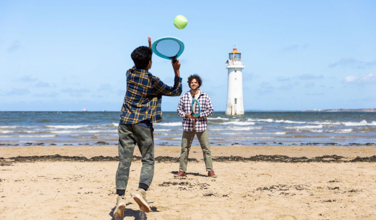 Two people playing tennis ona beach - there is a lighthouse in the background.