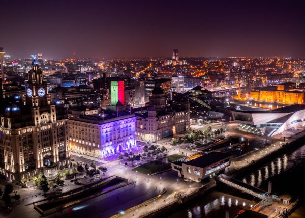 A drone shot of Liverpool's waterfront at night with the Three Graces buildings lit up.
